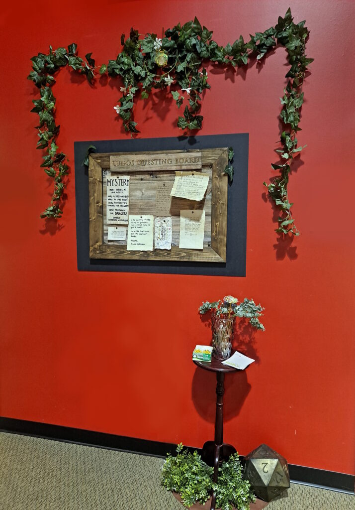 A wooden board labeled "Ludos Questing Board" with leafy vines drapped above. A glass vase filled with a sparkling translucent flower and soft leaves on a small wooden side table are below.