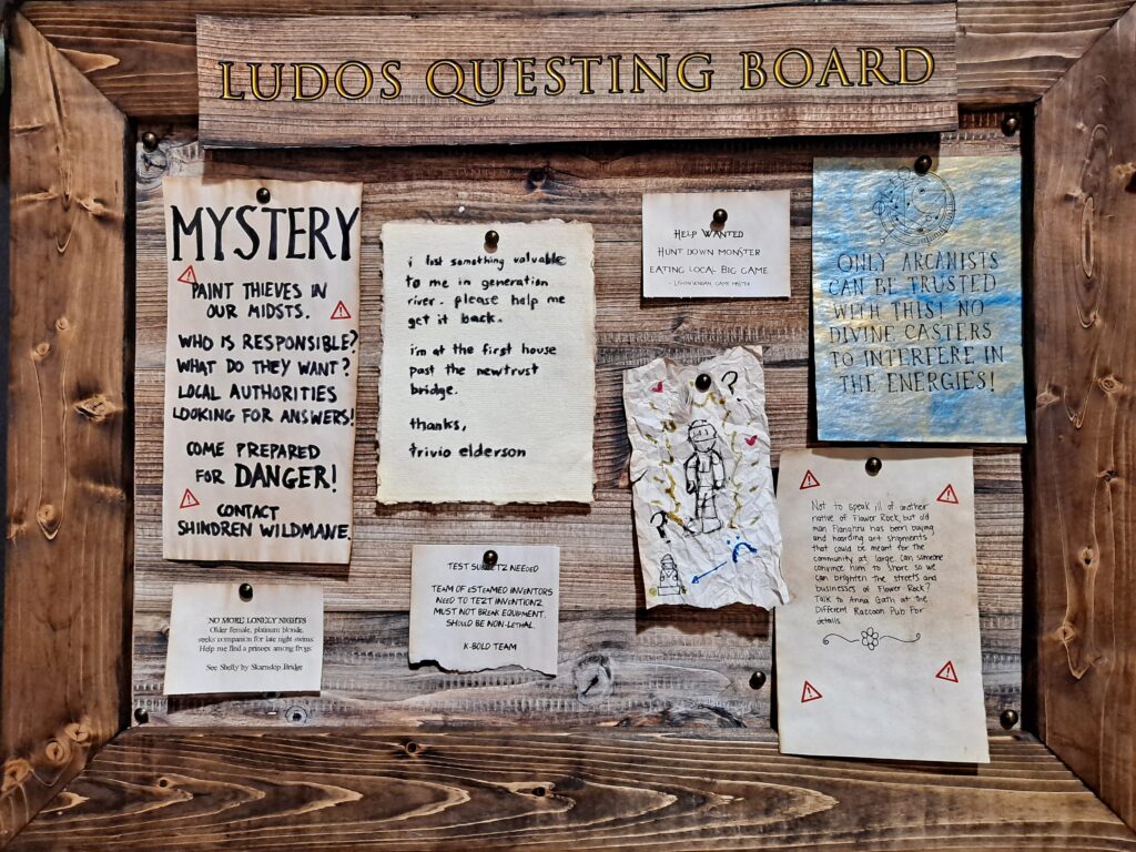 A close-up image of a wooden bulletin board labeled "Ludos Questing Board" filled with serveral parchment papers of various story hooks of varying sizes.