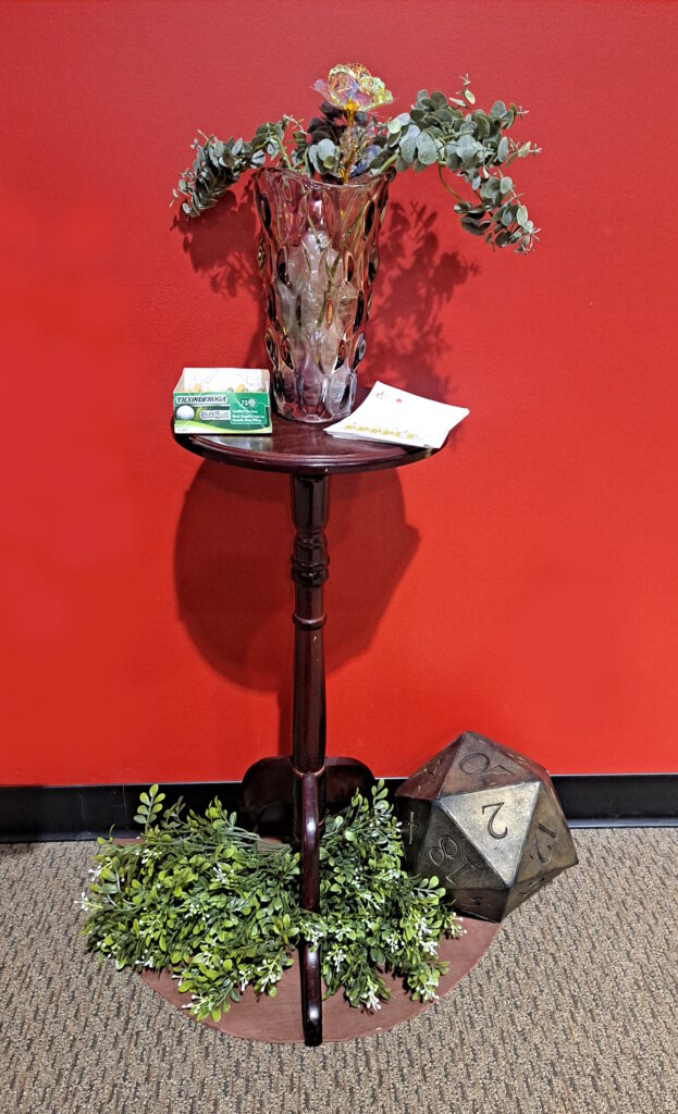 A close-up image of a Ludos Rockflower prop of a glass vase filled with a sparkling translucent flower and soft leaves on a small wooden side table with leafs decorating the bottom alongside a large d20 die.