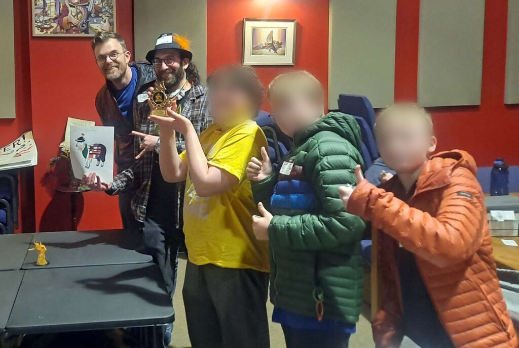 Photo of the five players that submitted the winning Garish Goat with their trophy. The faces of the younger adventures have been obscured.